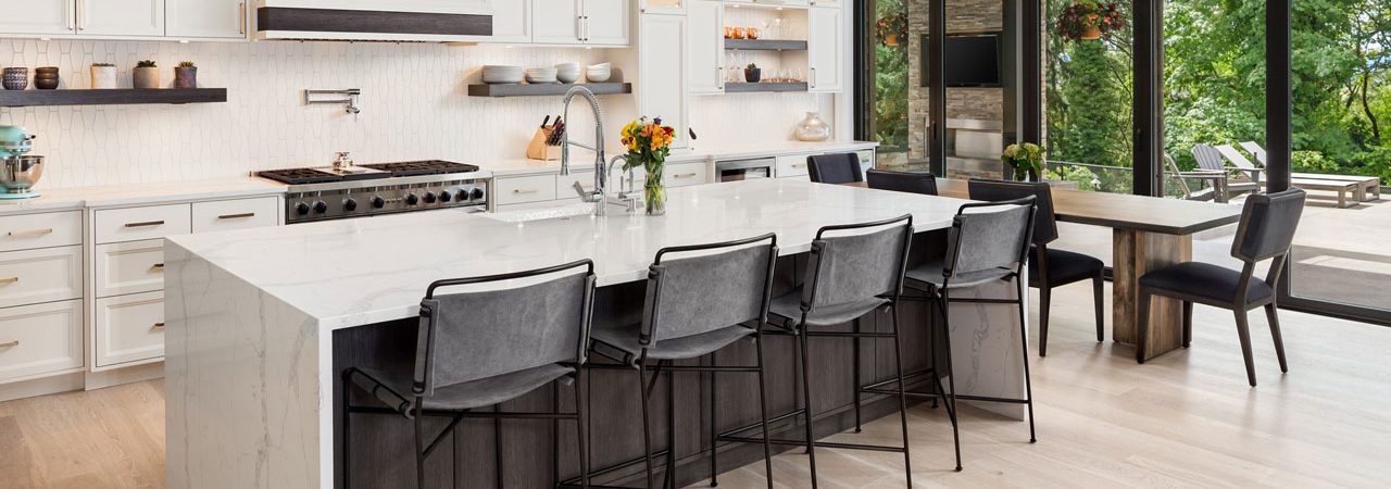 A remodeled kitchen with a marble island, a dining area, white cabinets and exhaust hood, and tiled backsplash